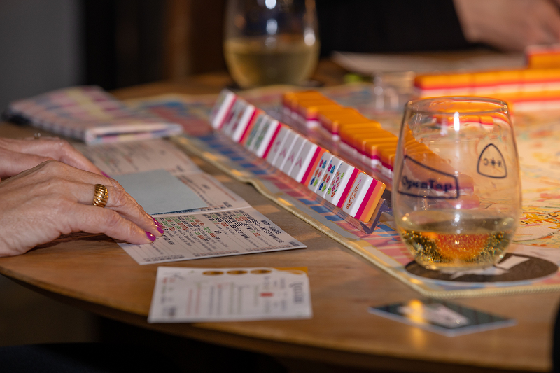 A close-up of hands holding cards next to a board game on a table, with two glasses of white wine nearby.
